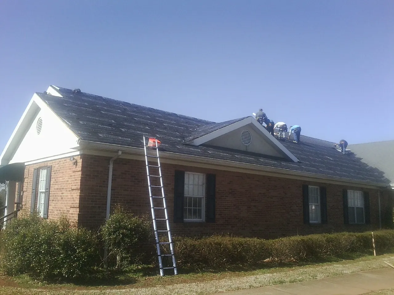 Crew tearing shingles off a brick ranch roof with a ladder set — Emergency Roof Repair work by our crew in Stryker