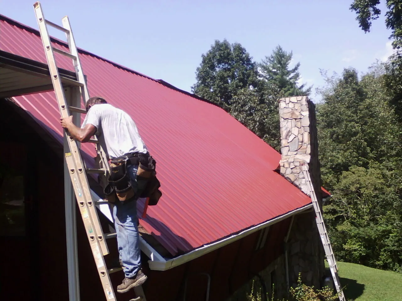 Roofer on a ladder beside a red metal roof and stone chimney — Soffit & Fascia Repair by Roofing Brothers in Stryker